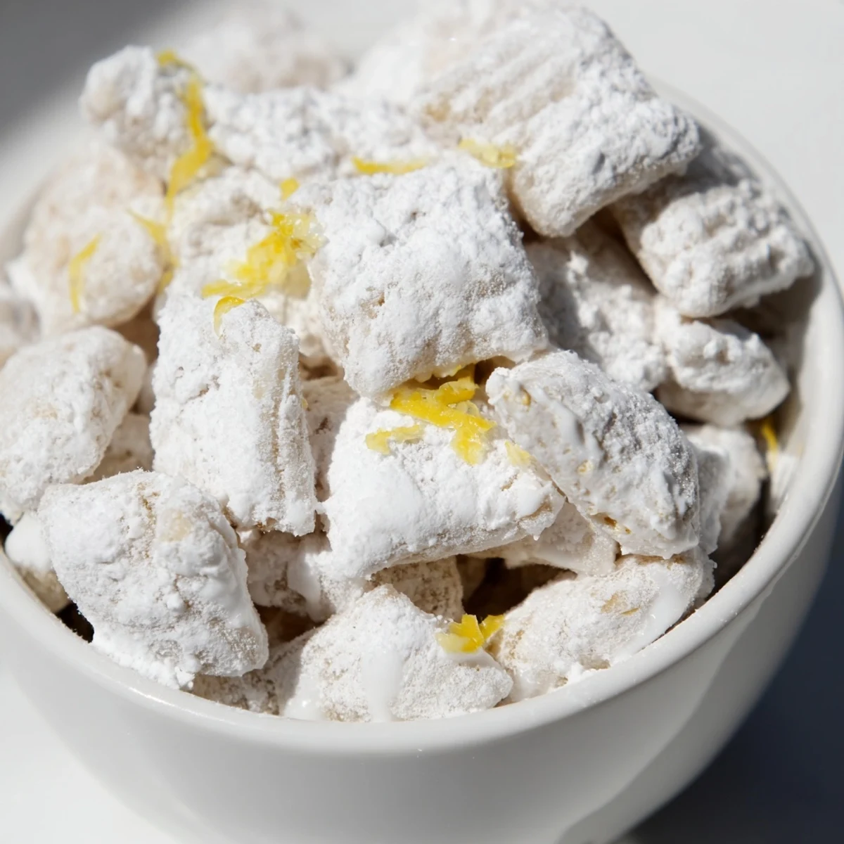 Golden lemonade puppy chow snack coated in white chocolate and powdered sugar spilling from a white bowl