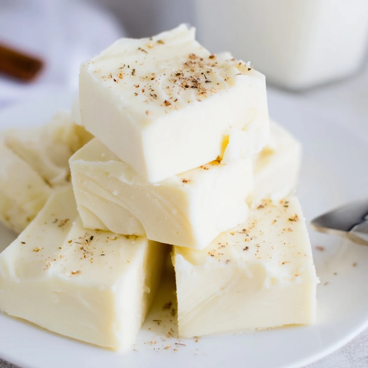Eggnog Fudge stacked on a plate, creamy white squares dusted with nutmeg