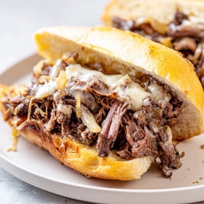 Tender slow-cooked French dip sandwiches with juicy beef overflowing from crusty bread alongside small bowls of savory beef broth for dipping